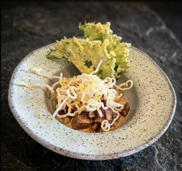 Cubos de atum em molho cremoso alaranjado, cobertos por harussame frito crocante e finalizados com folha de shissô em tempura, em bowl de cerâmica clara sobre pedra escura